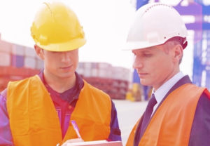 Two men in hard hats and orange vests looking at a clipboard.