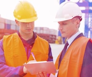 Two men in hard hats and orange vests looking at a clipboard.
